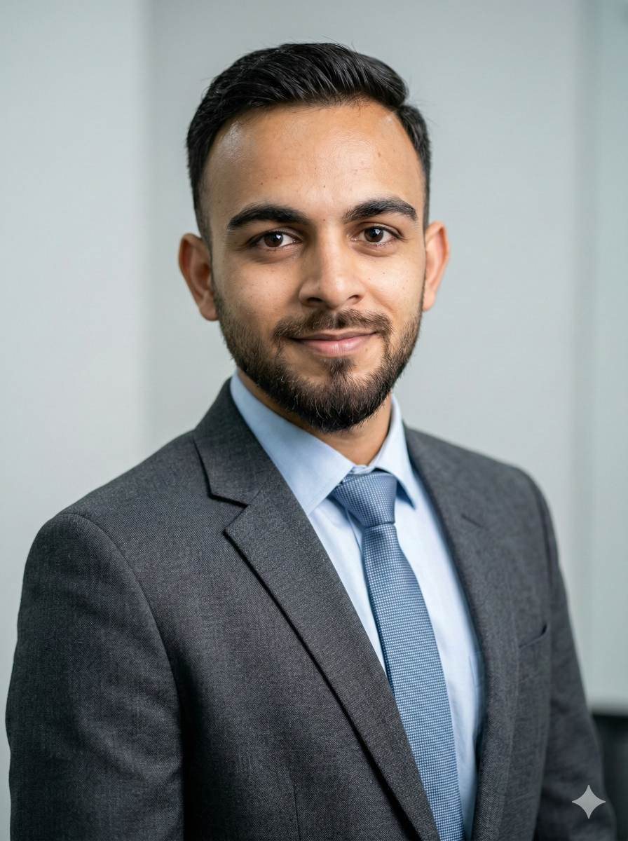 Muddassir Ali in a grey suit and blue tie, smiling, against a plain light wall